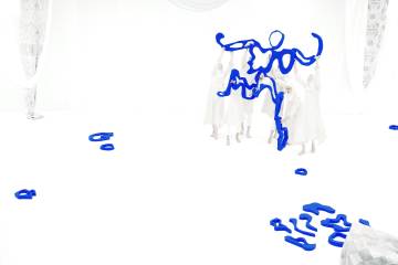 Against a white background, humans with pale faces wearing white robes hold up a vivid blue squiggly human-shaped figure with other blue squiggles scattered around the room
