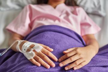 A girl lies in a hospital bed with an IV in her hand