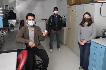 Team of three people in masks holding a small round device in a lab space