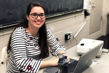 Woman in striped and floral top smiles while siting at a desk with a JHU water bottle and open laptop. A black board is behind her.