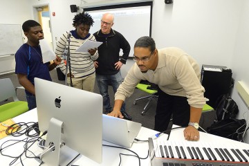 Thomas Dolby works with two young male singers at microphones while another man mixes sound on a laptop computer