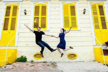 A man and woman hold hands and leap in the air in front of bright yellow row homes