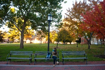 A female student reads a book on a campus quad bench in fall