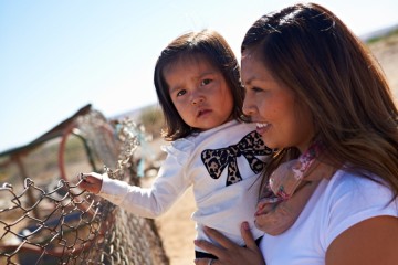 A close up image of a mother holding her toddler daughter up to a chain link fence