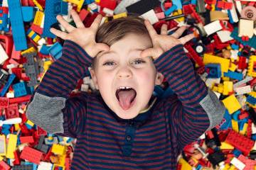 A happy little boy with dark blond hair and blue eyes lies in a pile of colorful plastic building blocks