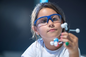 A little girl wearing safety goggles inspects a model of a molecule