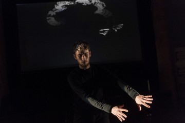 A man in front of a black screen waves his hand in front of a light while smokey swirls are projected on a screen behind him