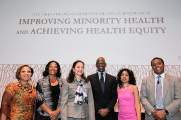 Six panelists at the Dean's Symposium pose for a photo in front of a screen with the talk title