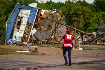 A trailer home lies on its side, having been ripped off its foundation, while a red cross volunteer looks on