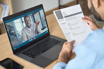 A man being interviewed appears on the laptop screen belonging to a hiring manager who is looking at the candidate’s resume