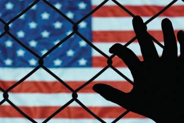 A hand reaching through a chain link fence toward the U.S. flag
