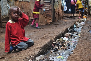 A young boy sits over an open sewer in the Kibera slum of Nairobi. The sewer is filled with trash and debris. The ground is packed dirt.