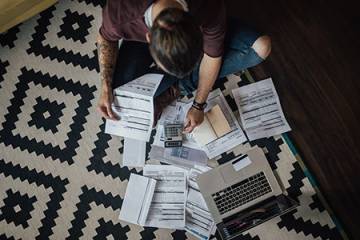 A person sits on the floor surrounded by bills, a calculator, and a laptop