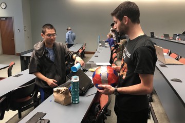 A student holds a soccer ball while another prepares a rolling object