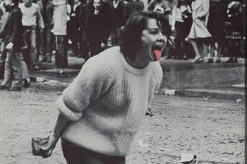 Excerpt of black and white Jorn photo shows a woman holding a rock and sticking out her tongue; her tongue is painted neon pink