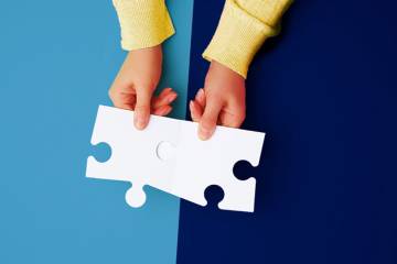 Top view of two hands joining white puzzle pieces over a background equally divided between dark blue and light blue