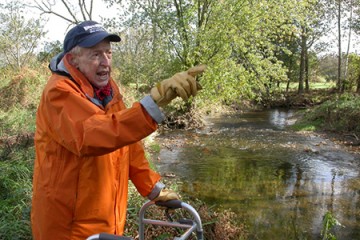 A man in a bright orange coat, a hat, and gloves, points to something outside the frame. There's a river and trees in the background