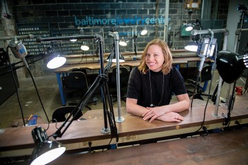 A woman sits at a work bench with her arms folded. There are adjustable lamps on all the workspaces, as well as various tools