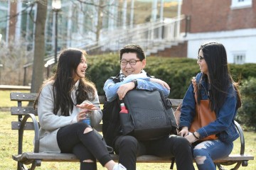 Two female students and a male student chat on a campus bench