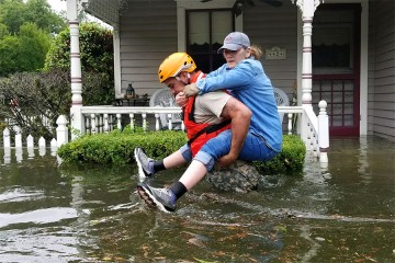 Man in yellow helmet, orange vest carries woman piggyback style amid waist-deep floodwaters in Houston