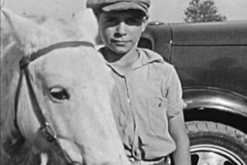 A black and white photo shows a boy in a newsy cap holding the reins of a white horse