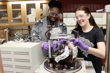 Two women wearing safety goggles attach foil to a piece of lab equipment