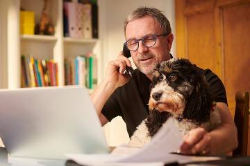 Man working at computer on table at home with dog on his lap