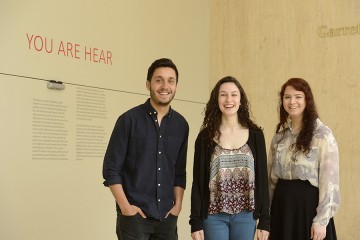 Three students pose in from of 'Your Are Hear' exhibition title in red letters on wall