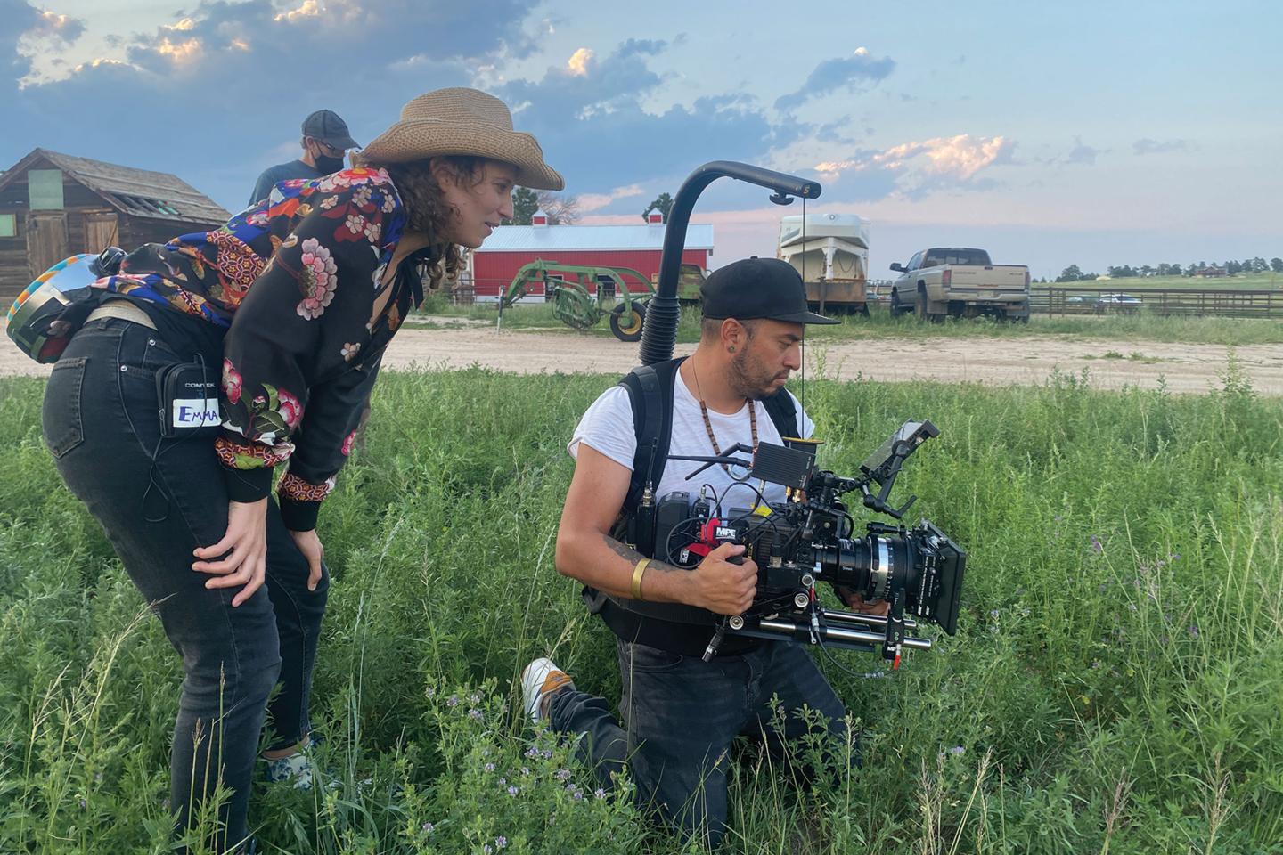 Emma Needell, wearing a cowboy hat, looks on as a someone holding a video camera films a scene