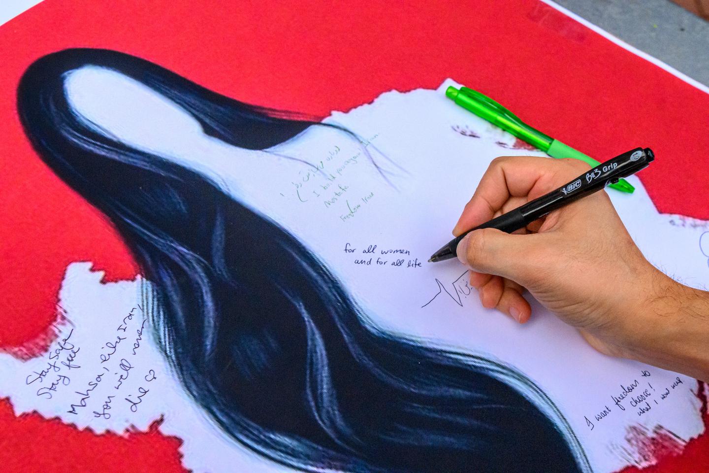 An individual writes a message on a person depicting a faceless woman with long, black hair over a silhouette of Iran laid against a red background