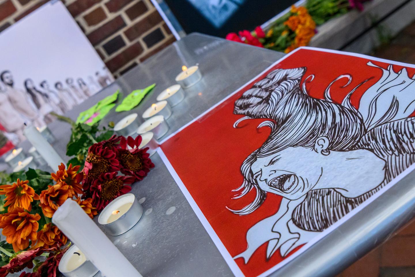 A poster depicts a woman with long hair screaming; it sits on a table next to a few burning candles and a bouquet of orange and red flowers