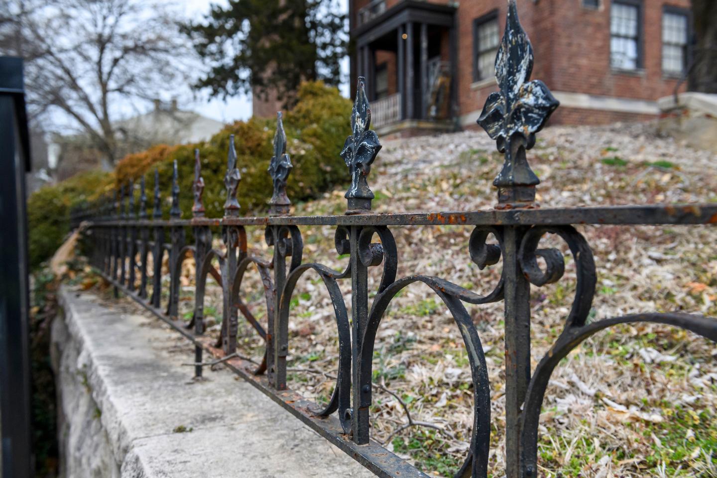 A close-up view of a black, rusted wrought iron fence with a brick building in the background