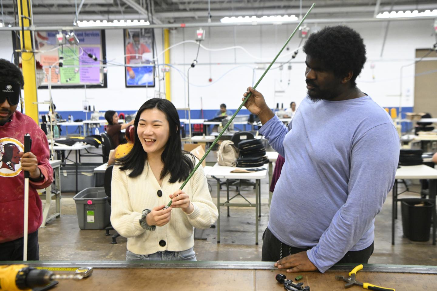 Students work in partnership with blind individuals to make canes. A student in a white sweater holds up a cane made from a green tomato stake while a blind associate at Blind Industries and Services of Maryland learns how to make the cane.