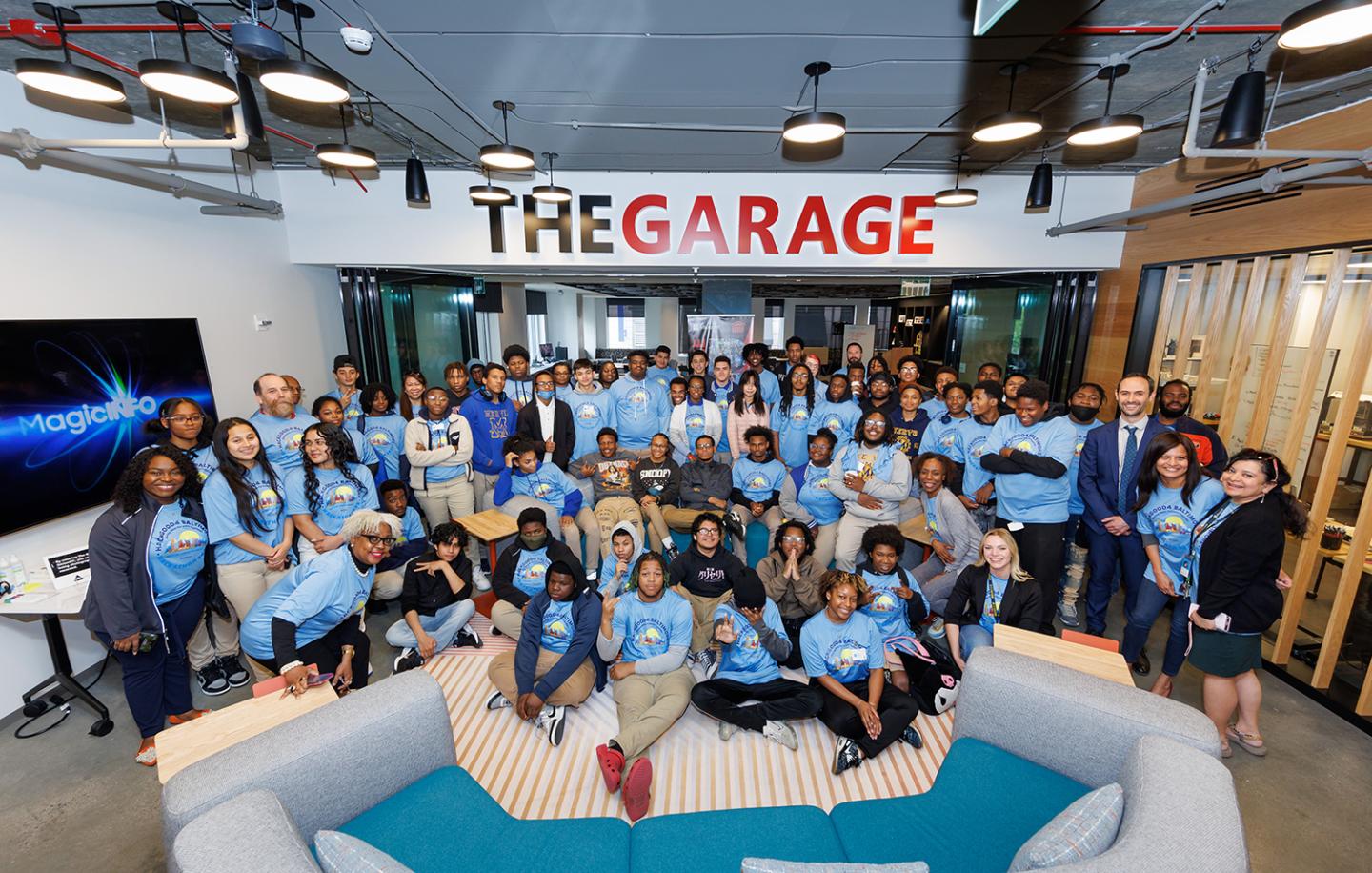 A large group, most wearing light blue t-shirts, poses for a photo on a large sign that reads 'The Garage'