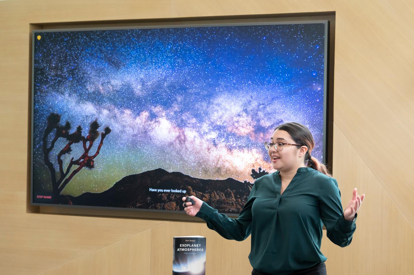 A young woman in a dark green blouse stands in front of a large display screen with colorful image of the night sky and stars