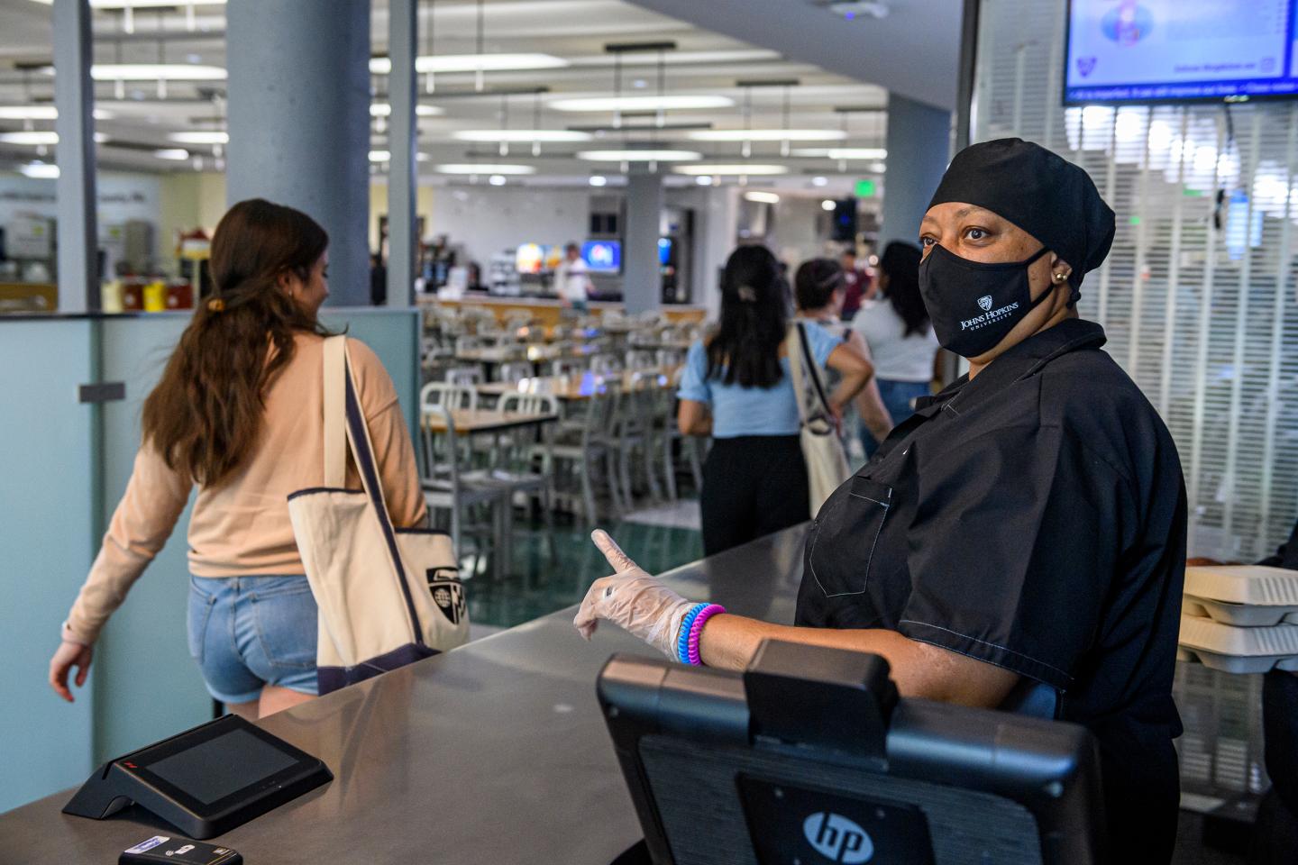 A woman in a black hat, black mask, and black shirt stands at a cash register as students file past her into a dining area