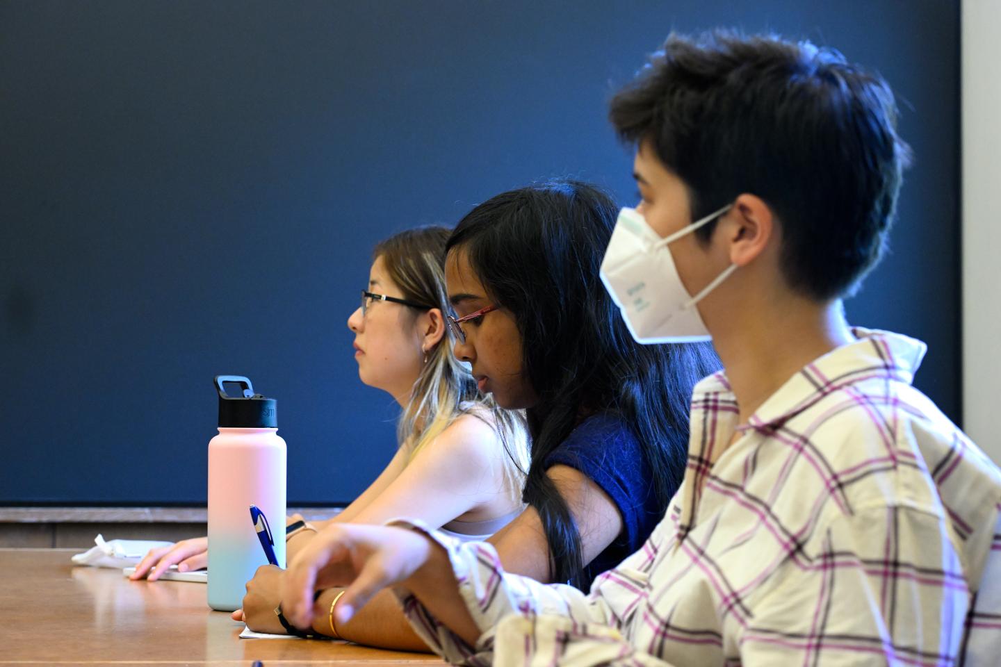 Three students sit in a classroom, attentive; one is wearing a white mask; a blackboard is in the background