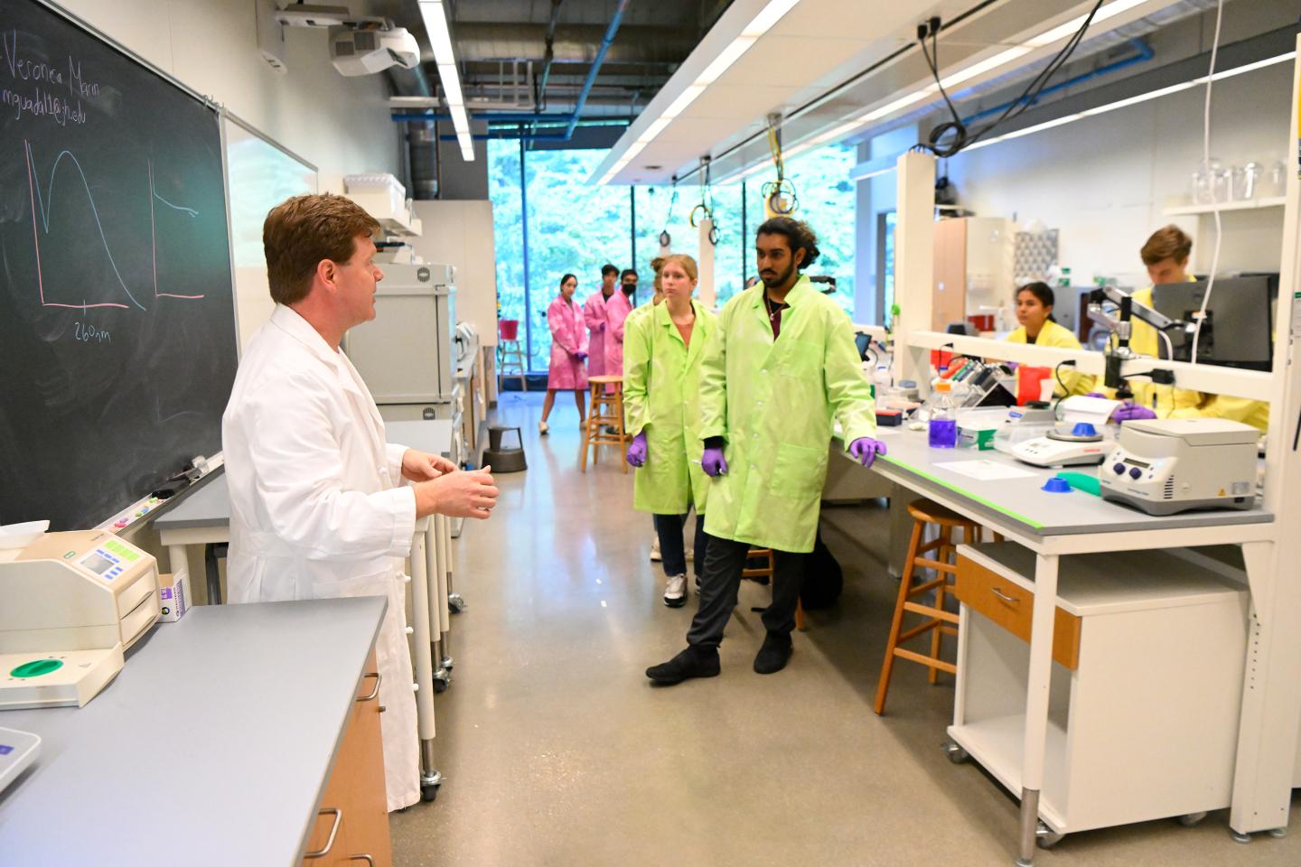 An instructor in a white lab coat addresses a class in a lab setting