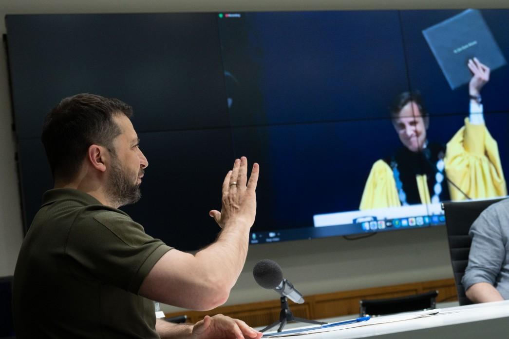 President Volodymyr Zelenskyy waves as he is presented with an honorary degree by Johns Hopkins University President Ron Daniels, visible on a screen in the background