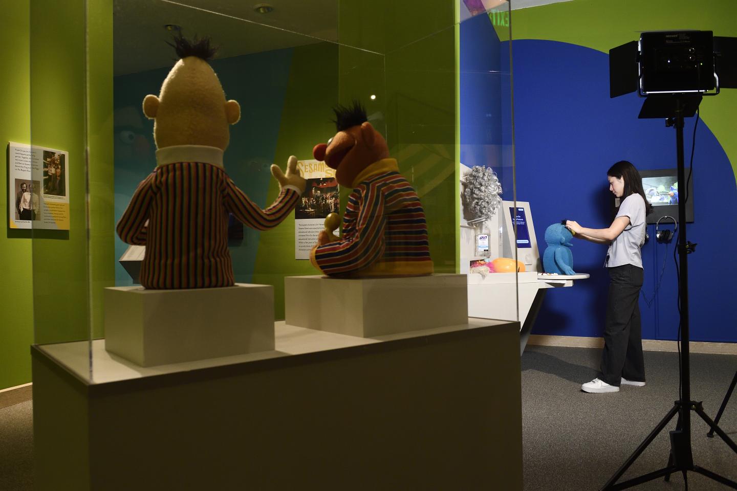 A display case with Burt and Ernie puppets is photographed from the back. Beyond the case, a young adult makes their own puppet.