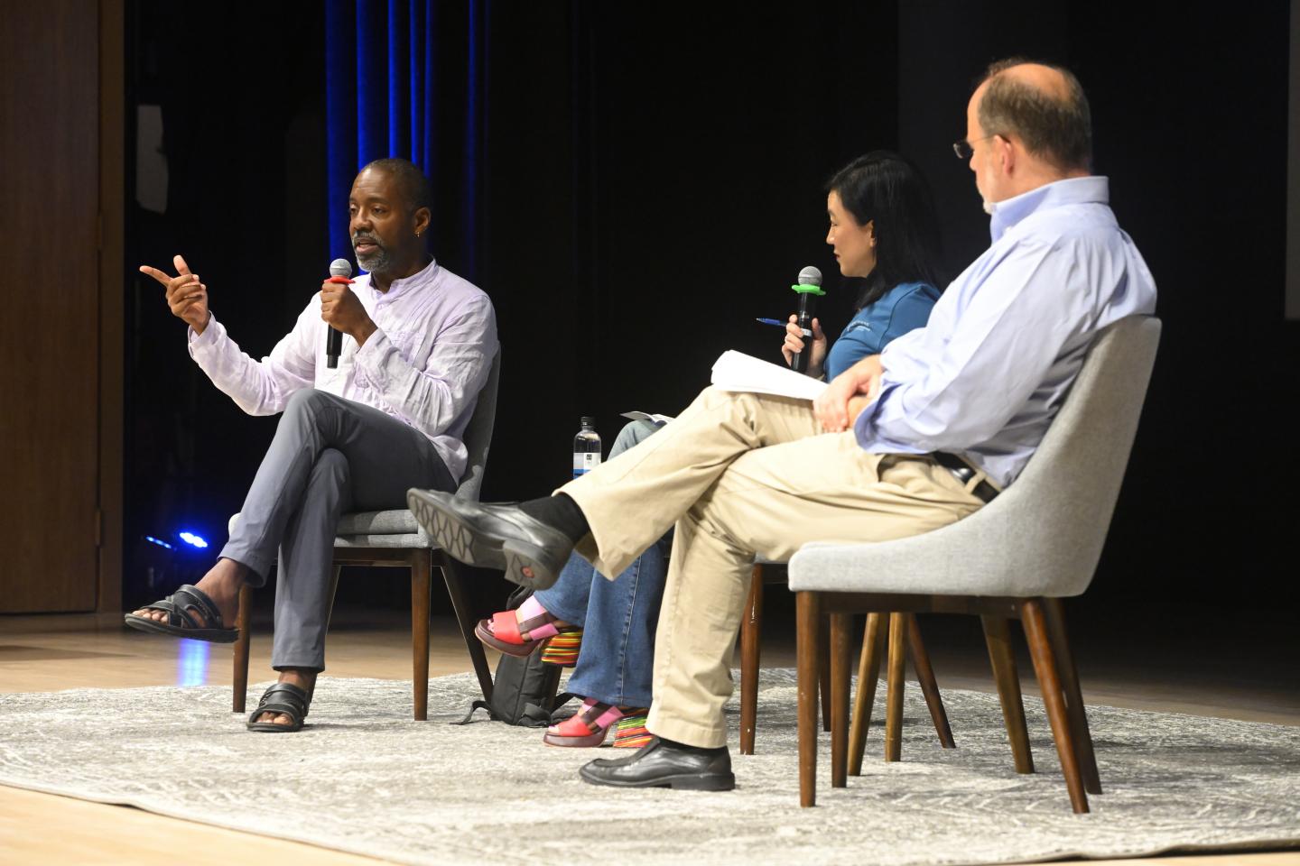 Three people sit on chairs on a stage, having a discussion. They are holding microphones.