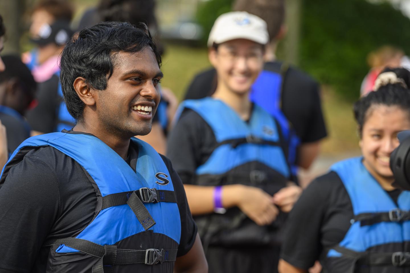 A student smiles while standing in a blue life vest. Behind him, two others do the same.