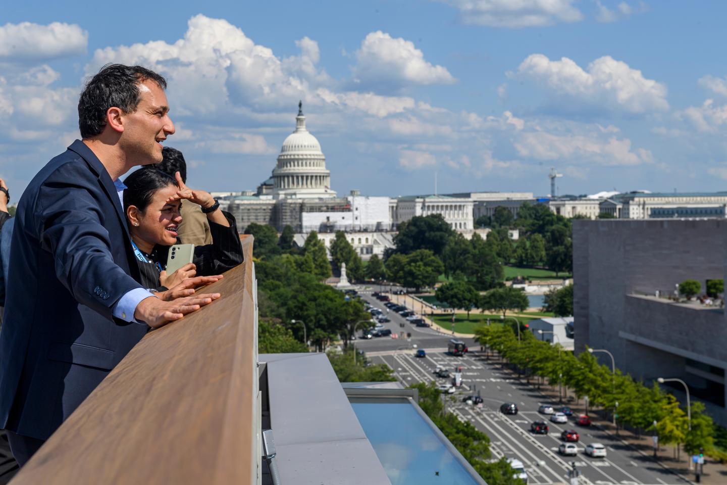 Two people stand on the roof and look out on Pennsylvania Avenue; the U.S. Capitol dome is prominently visible in the background