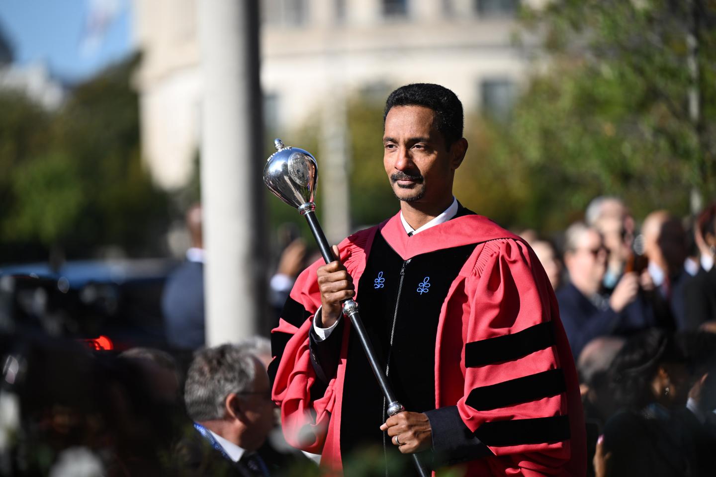 A man in red regalia carries a silver mace