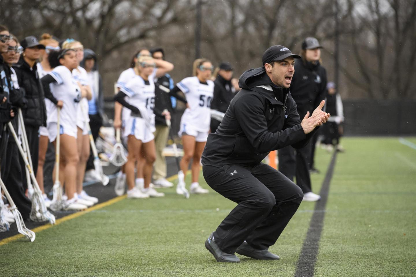 Coach Tim McCormack crouches and claps his hands on the sidelines of a women's lacrosse game.
