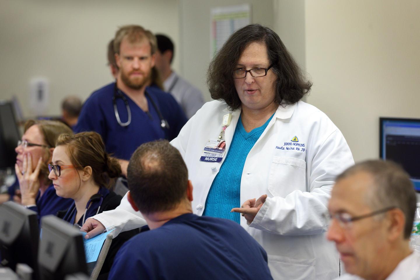 A nurse in a white coat speaks to a group of nursing student in dark blue scrubs