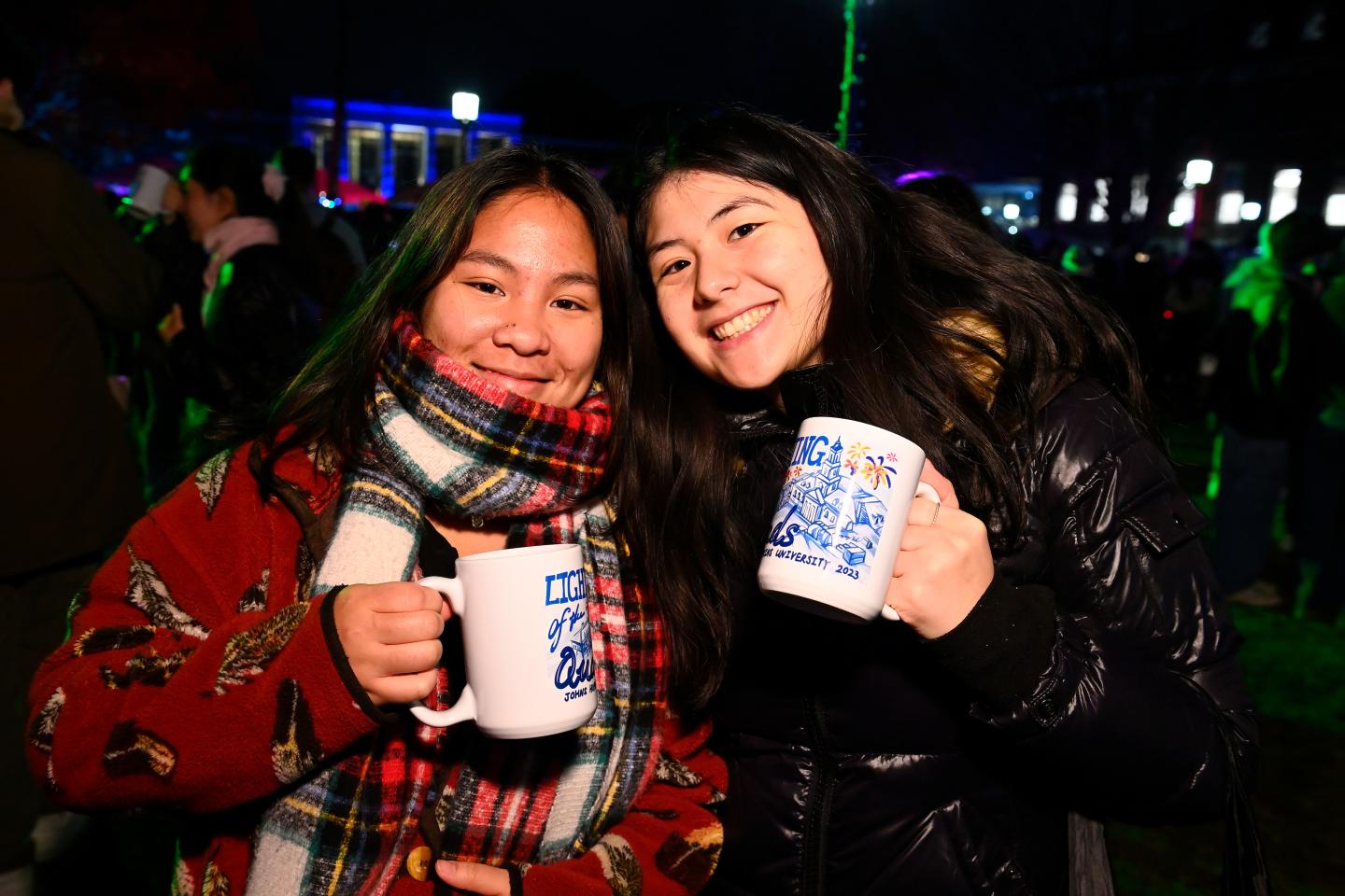 Two female students smile while holding their limited edition 2023 lighting of the quads mugs 