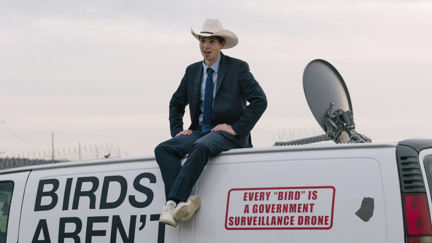 A young man wearing a white cowboy hat sits atop a white van; on the van is written 