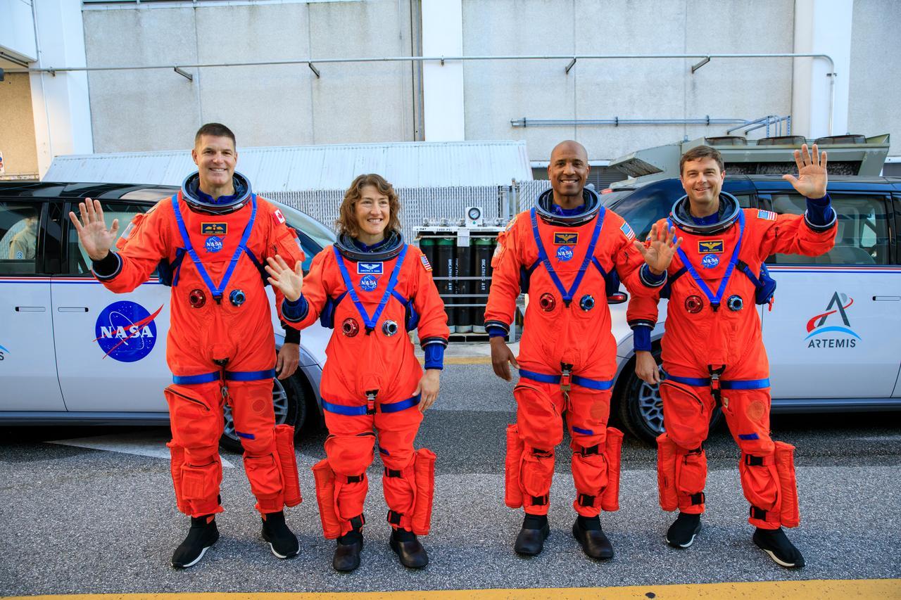 Four astronauts stand and wave while wearing orange jumpsuits