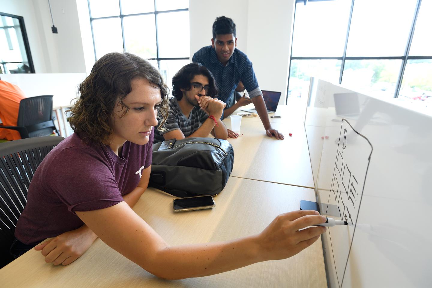 A person writes on a whiteboard while two others look on